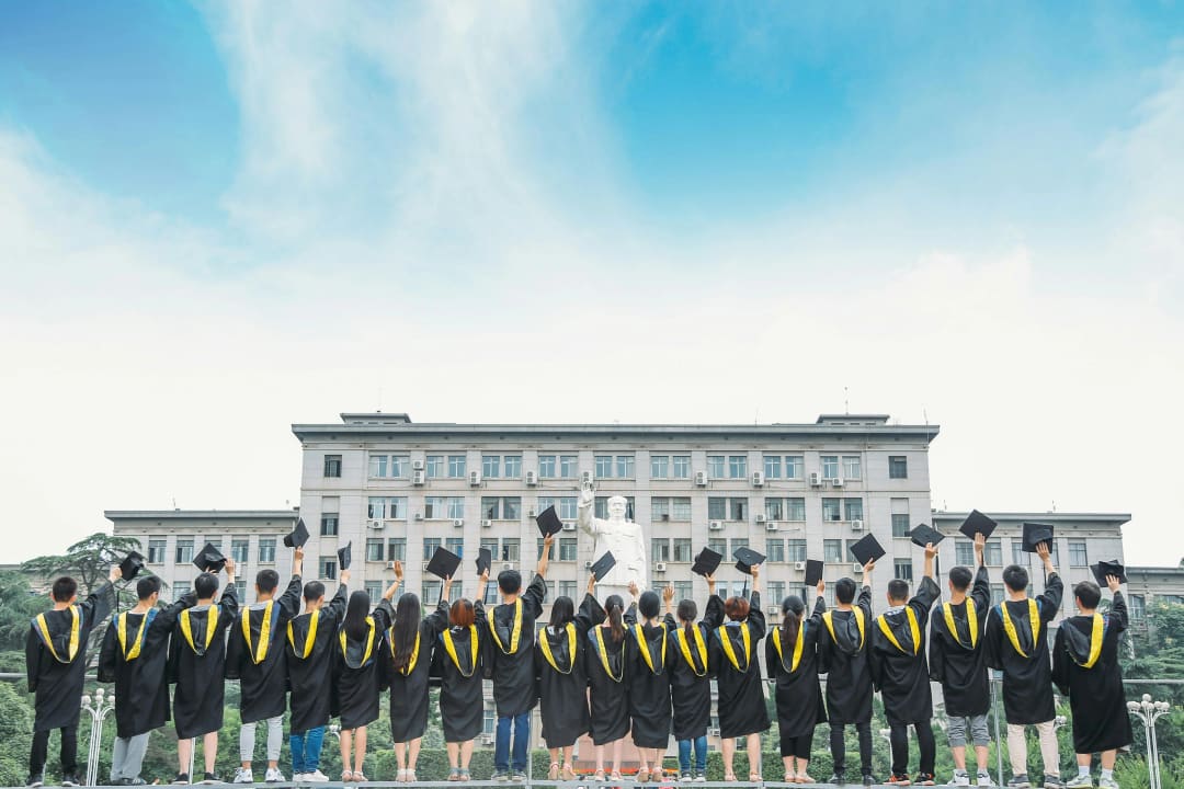 Students in gowns, seen from behind, raising their graduation caps in front of their university building.